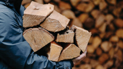 Unrecognizable man carries heap of wood for making fire dressed in jacket. Faceless male carries firewood into house