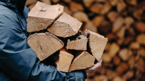 Unrecognizable man carries heap of wood for making fire dressed in jacket. Faceless male carries firewood into house